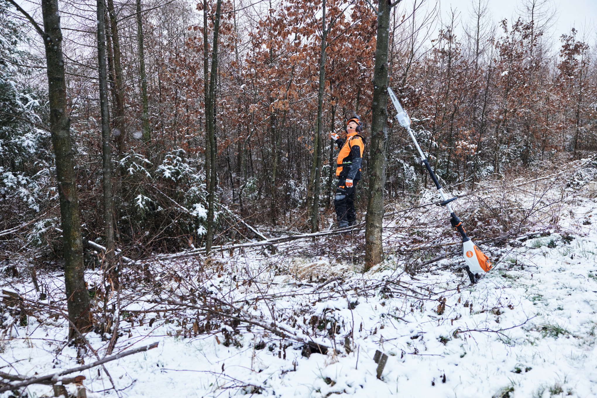 Um homem a trabalhar com equipamento de proteção individual numa floresta coberta de neve, em primeiro plano, uma podadora em altura a bateria STIHL HTA 150 encostada a um tronco de árvore