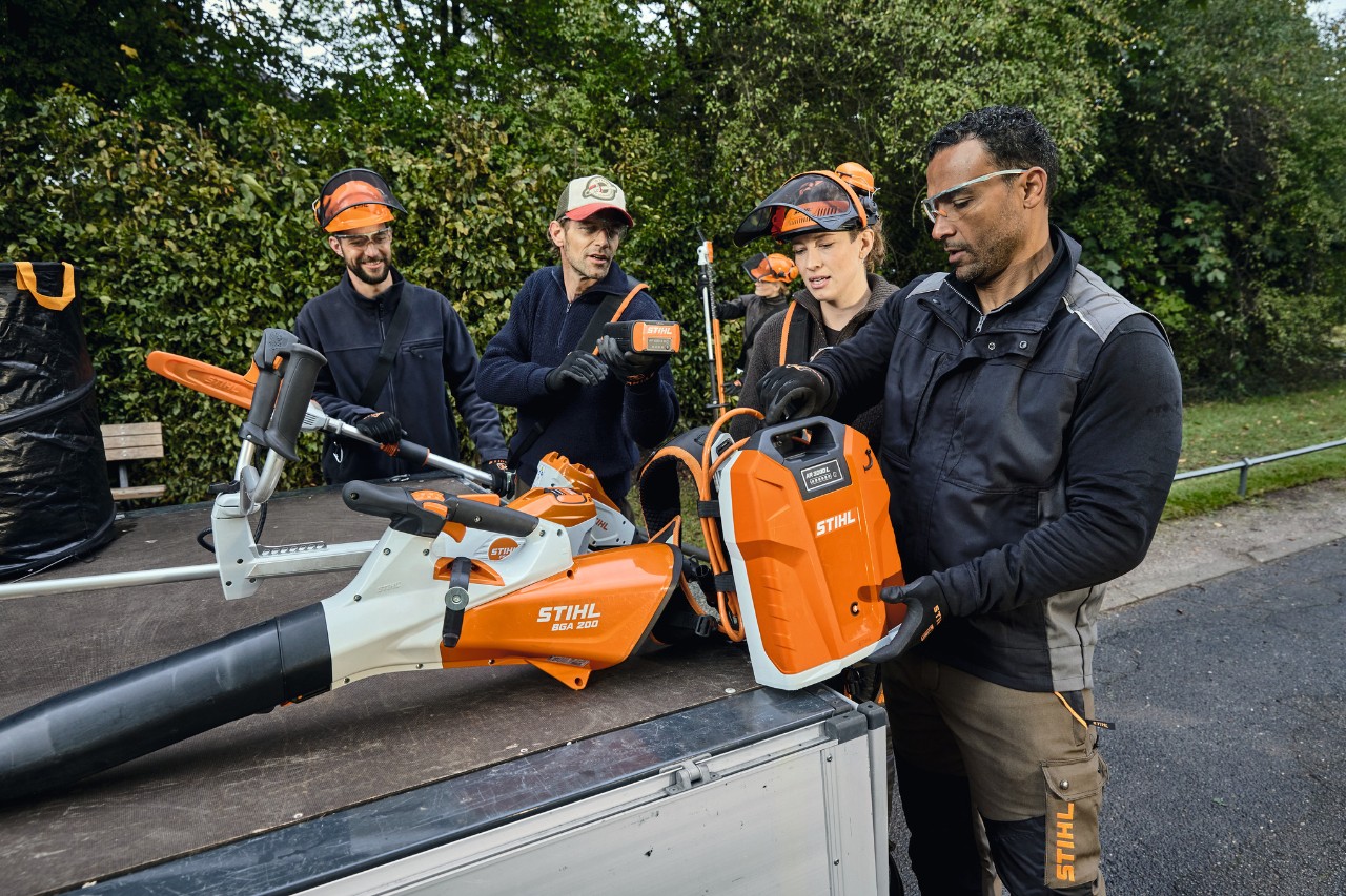 Três homens e uma mulher com equipamento de proteção STIHL na superfície de carga de um veículo com máquinas STIHL, como, por exemplo, o soprador de folhas a bateria STIHL BGA 200 e a bateria de mochila STIHL AR 2000 L 