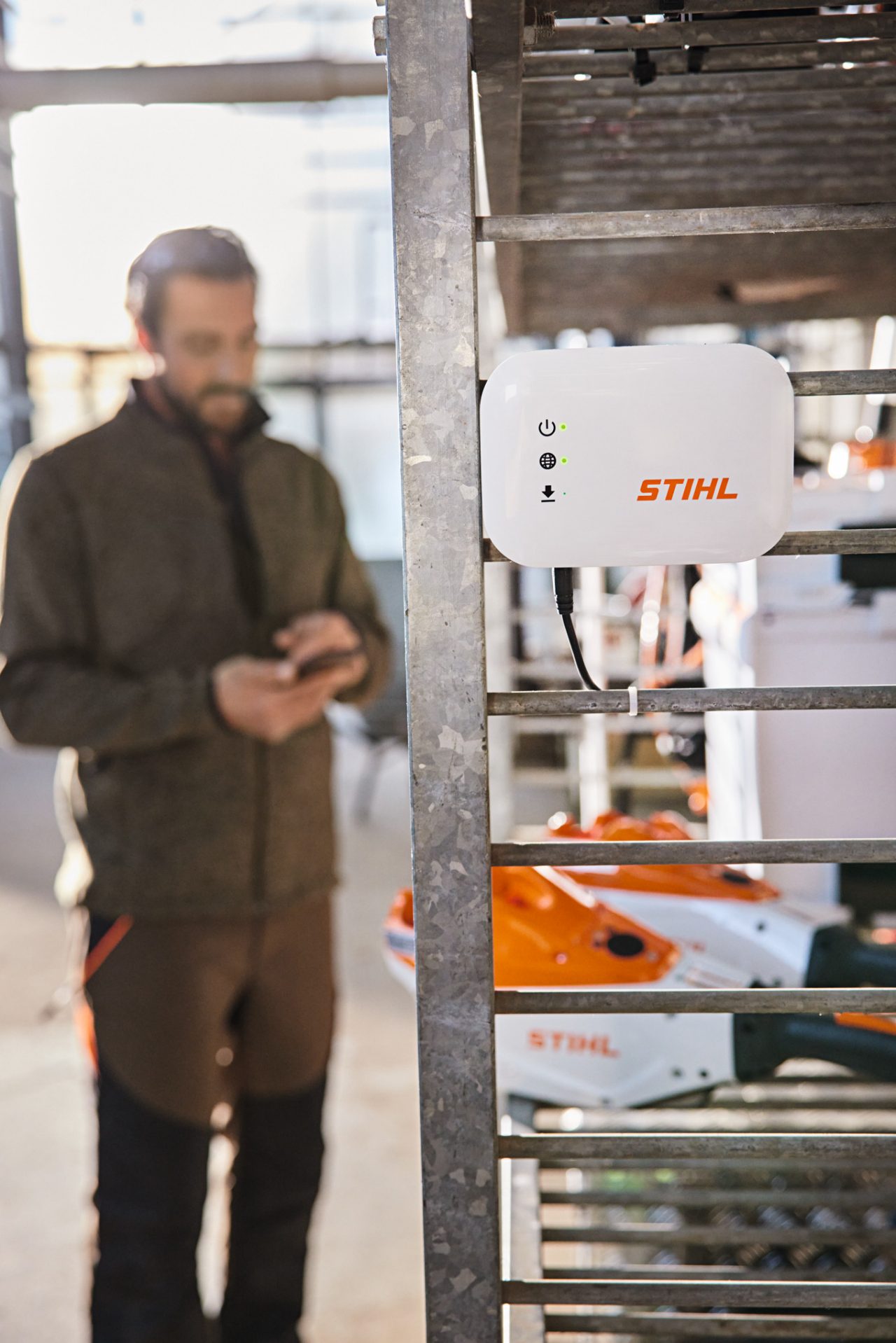 Uma STIHL connected Box pendurada numa estante de oficina. Em segundo plano, um homem interage com o seu smartphone. 