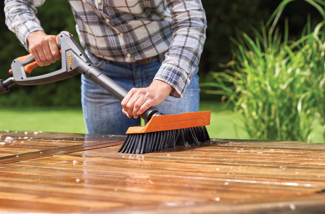 Uma mulher a limpar uma mesa de madeira para jardins com uma máquina de limpeza de alta pressão STIHL RE 130 Plus com escova de lavagem
