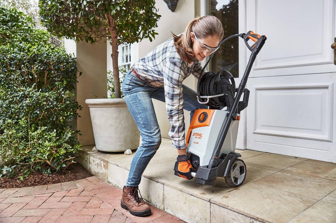 Uma mulher a levantar uma máquina de limpeza de alta pressão STIHL RE 130 Plus num caminho de tijolo