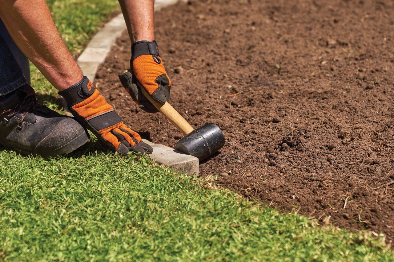 Como plantar perenes: uma pessoa de luvas com um maço para criar uma borda de tijolos num canteiro de jardim