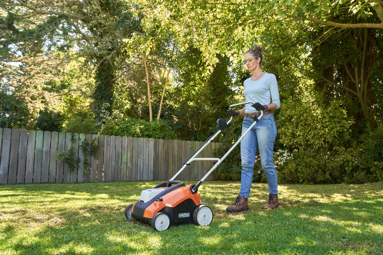 Uma mulher ventila o relvado no espaço verde com o arejador de relva a bateria STIHL RLA 240, equipada com óculos de proteção e luvas de trabalho.