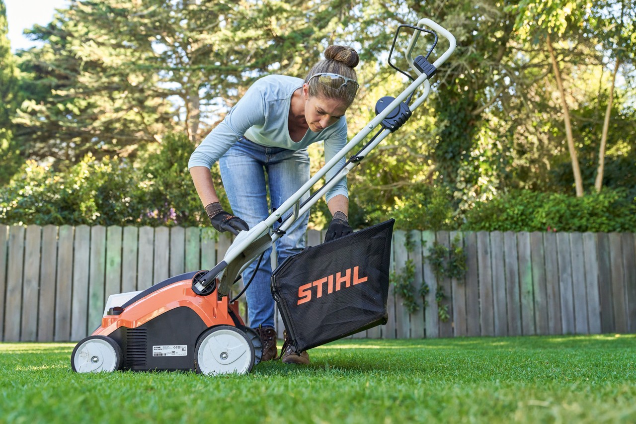 Uma mulher pendura o recolhedor do arejador de relva a bateria STIHL RLA 240 na máquina antes de arejar a relva