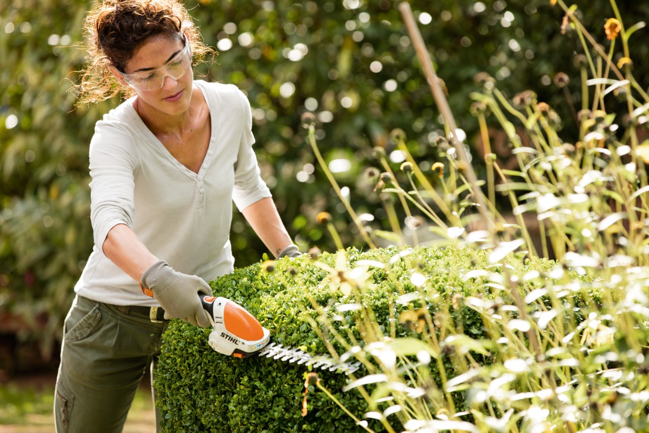 Uma mulher com óculos de segurança e luvas mostra como aparar sebes com a minipodadora a bateria STIHL HSA 25
