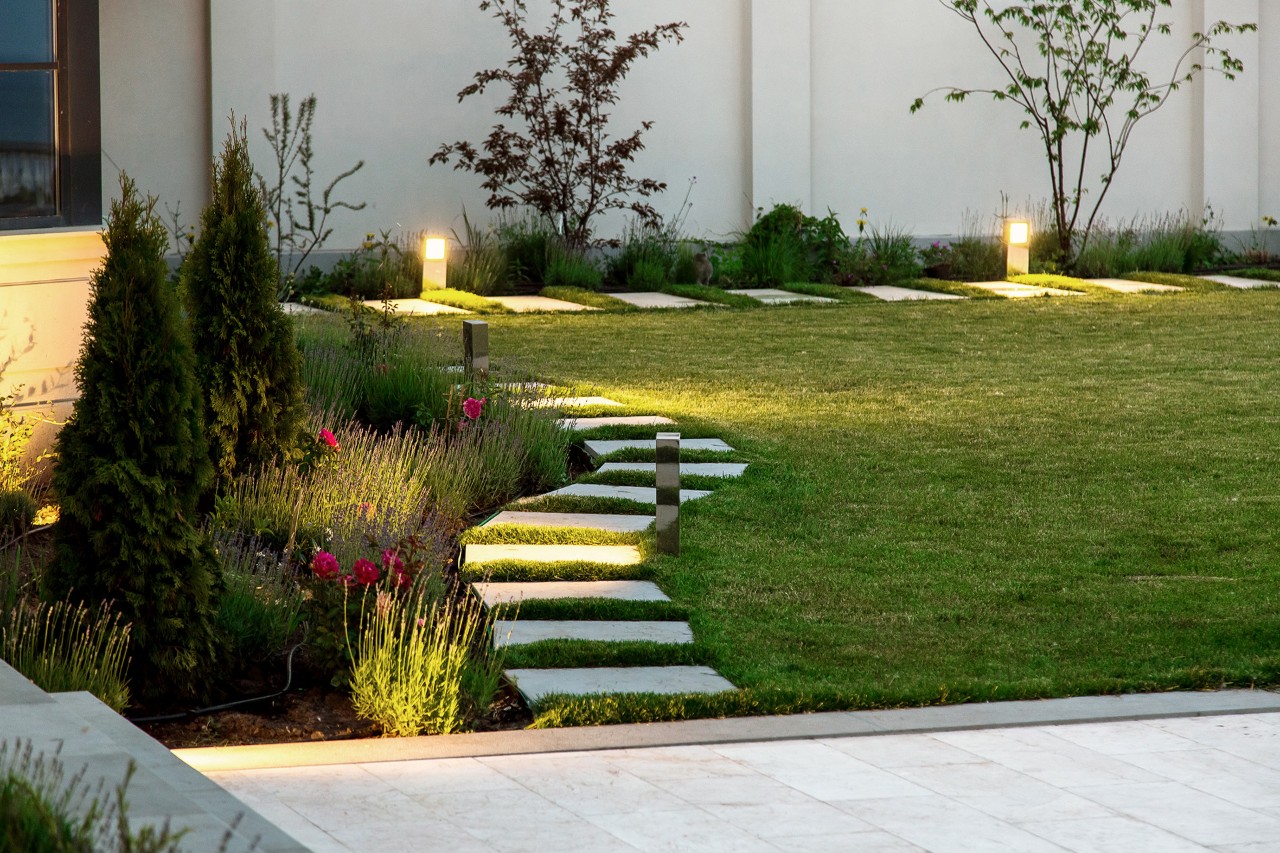 Jardim pequeno à noite, canteiro de flores, relvado verde, carreiro de lajes quadradas de mármore, iluminação decorativa do chão.