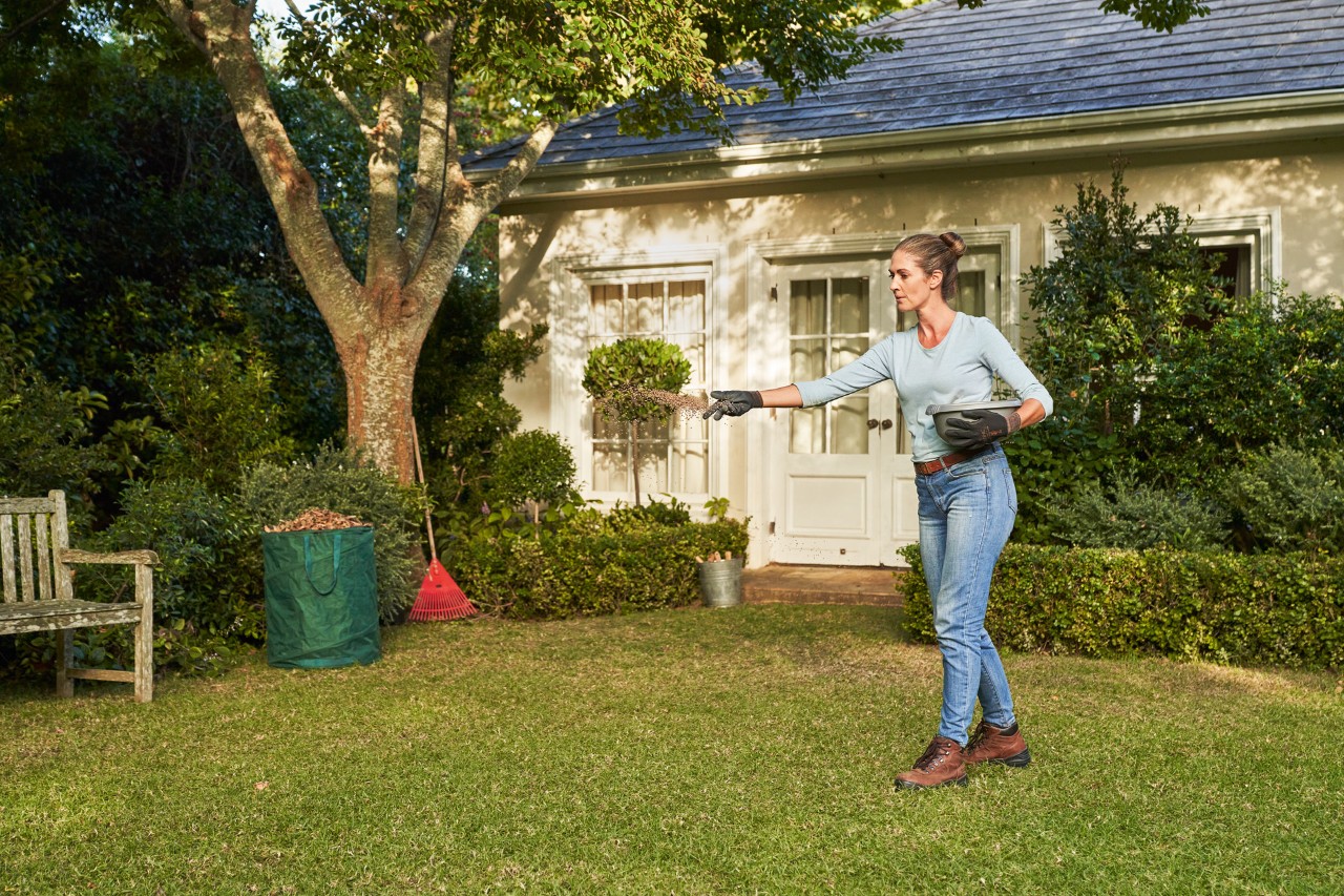 Uma mulher com luvas STIHL FUNCTION SENSOTOUCH espalha cal de jardim no relvado; em segundo plano, arbustos e uma casa