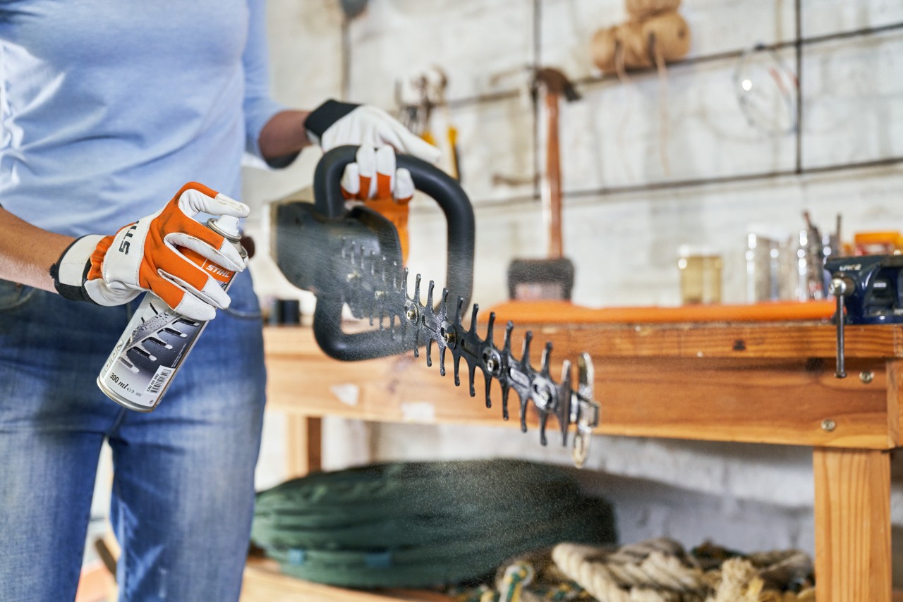Uma mulher com luvas faz a manutenção do corta-sebes a bateria STIHL HSA 56 ao pulverizar solvente de resina na lâmina