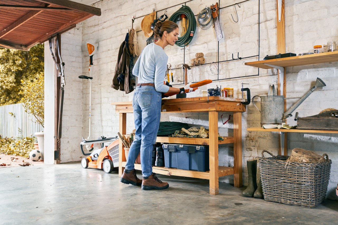 Uma mulher equipada diante de uma bancada numa garagem, procede à manutenção do corta-sebes a bateria STIHL HSA 56