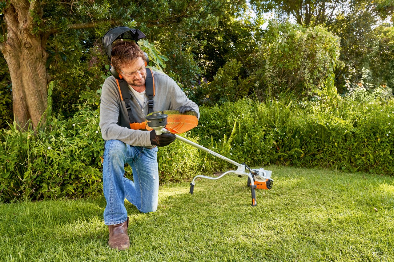 Um homem com equipamento de proteção a demonstrar como substituir uma cabeça de motorroçadora num jardim 