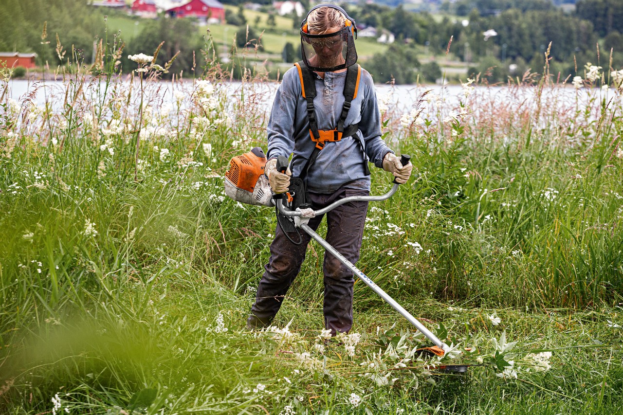 Uma mulher com proteção facial demonstra como usar uma motorroçadora STIHL com erva alta, ao lado de um lago