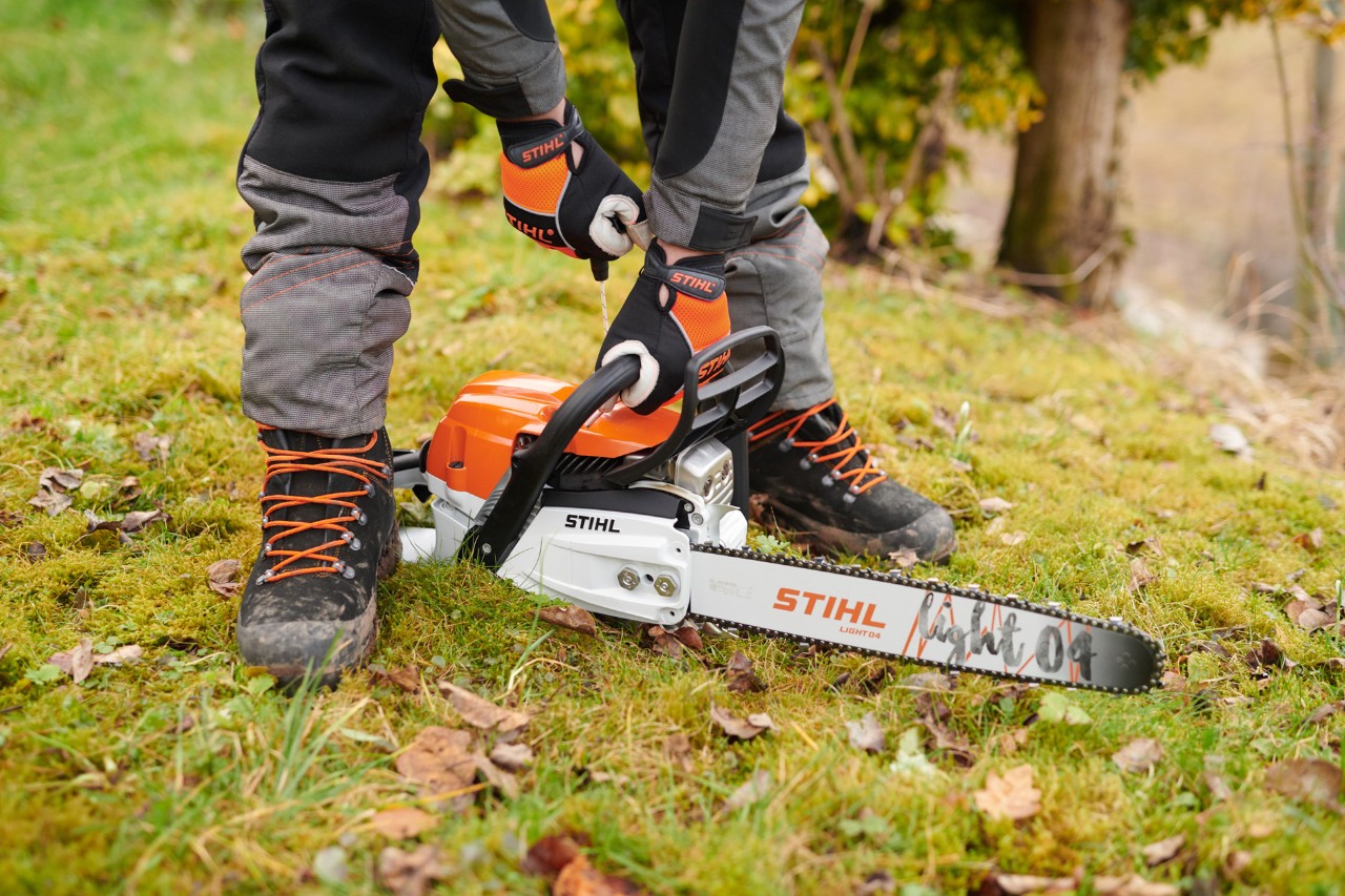 Alguém com luvas mostra como ligar uma motosserra STIHL MS 261 C-M a gasolina com guia Light 04, no chão