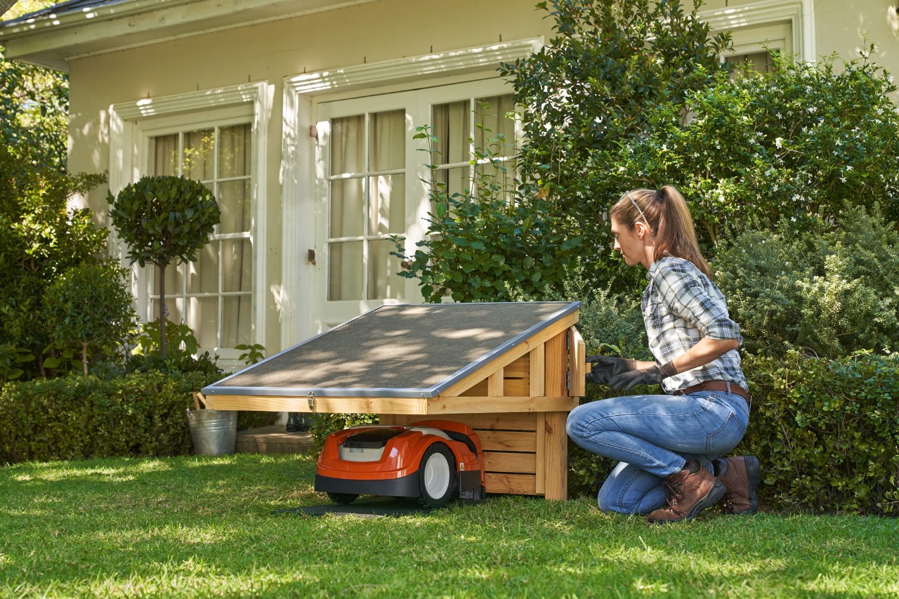 Uma mulher ajoelhada no relvado junto ao robot corta-relva STIHL iMOW® estacionado abre o compartimento de arrumação
