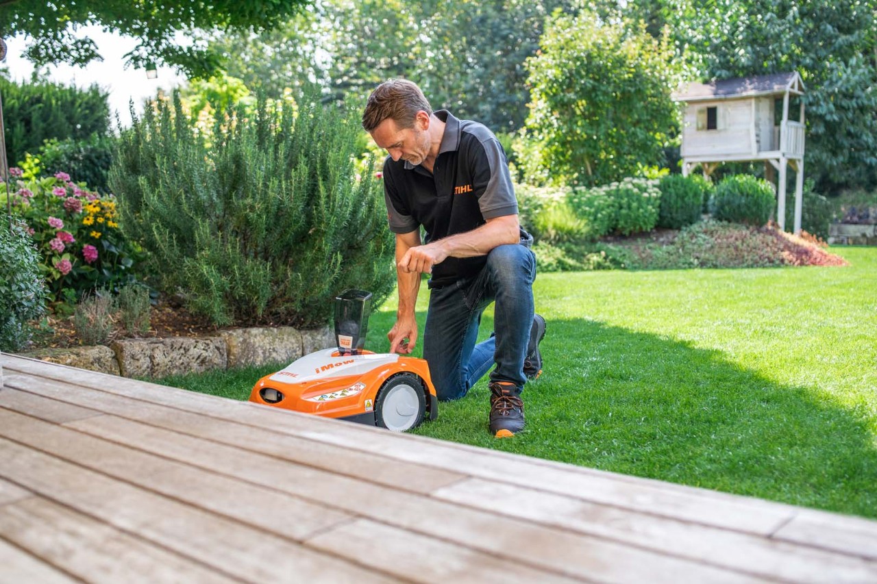 Um homem ajoelhado num relvado após a limpeza do robot corta-relva STIHL iMOW®, num jardim com arbustos e um alpendre