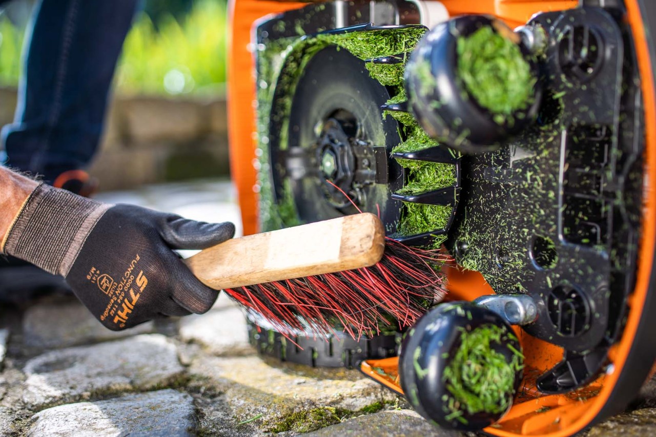 Alguém com luvas e uma escova procede à limpeza do robot corta-relva STIHL iMOW®, na plataforma de corte cheia de relva