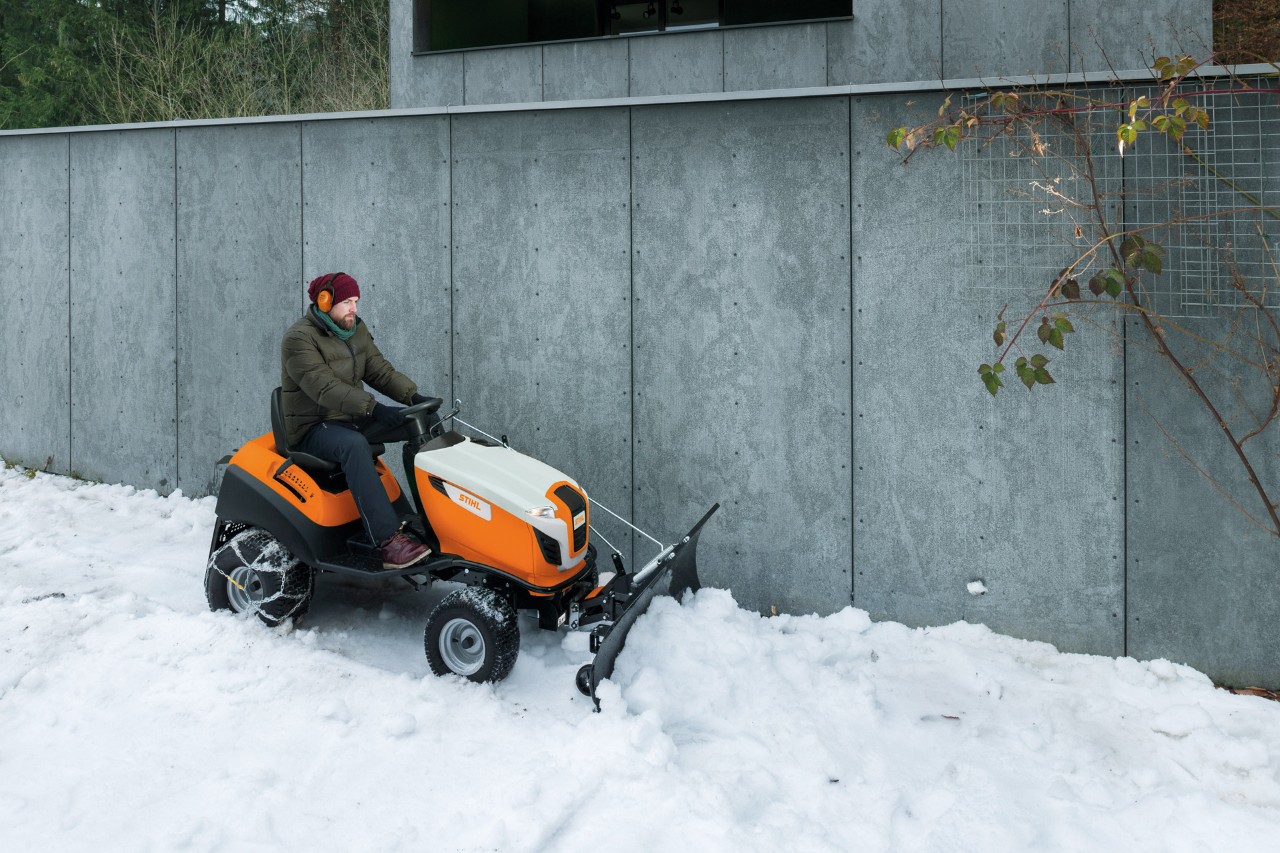 Um homem sentado num trator corta-relva com uma lâmina de neve limpa neve em frente a um muro 