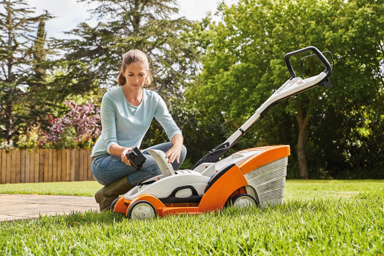 Uma mulher ajoelhada ao lado de um corta-relva STIHL RMA 339 C e a remover a bateria
