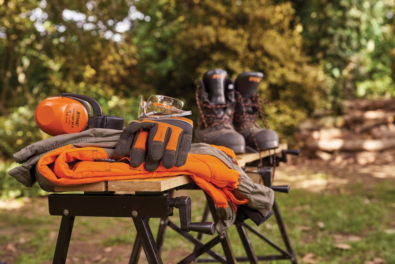 Proteção auditiva STIHL, botas, óculos de proteção e luvas de proteção numa bancada de trabalho num espaço verde