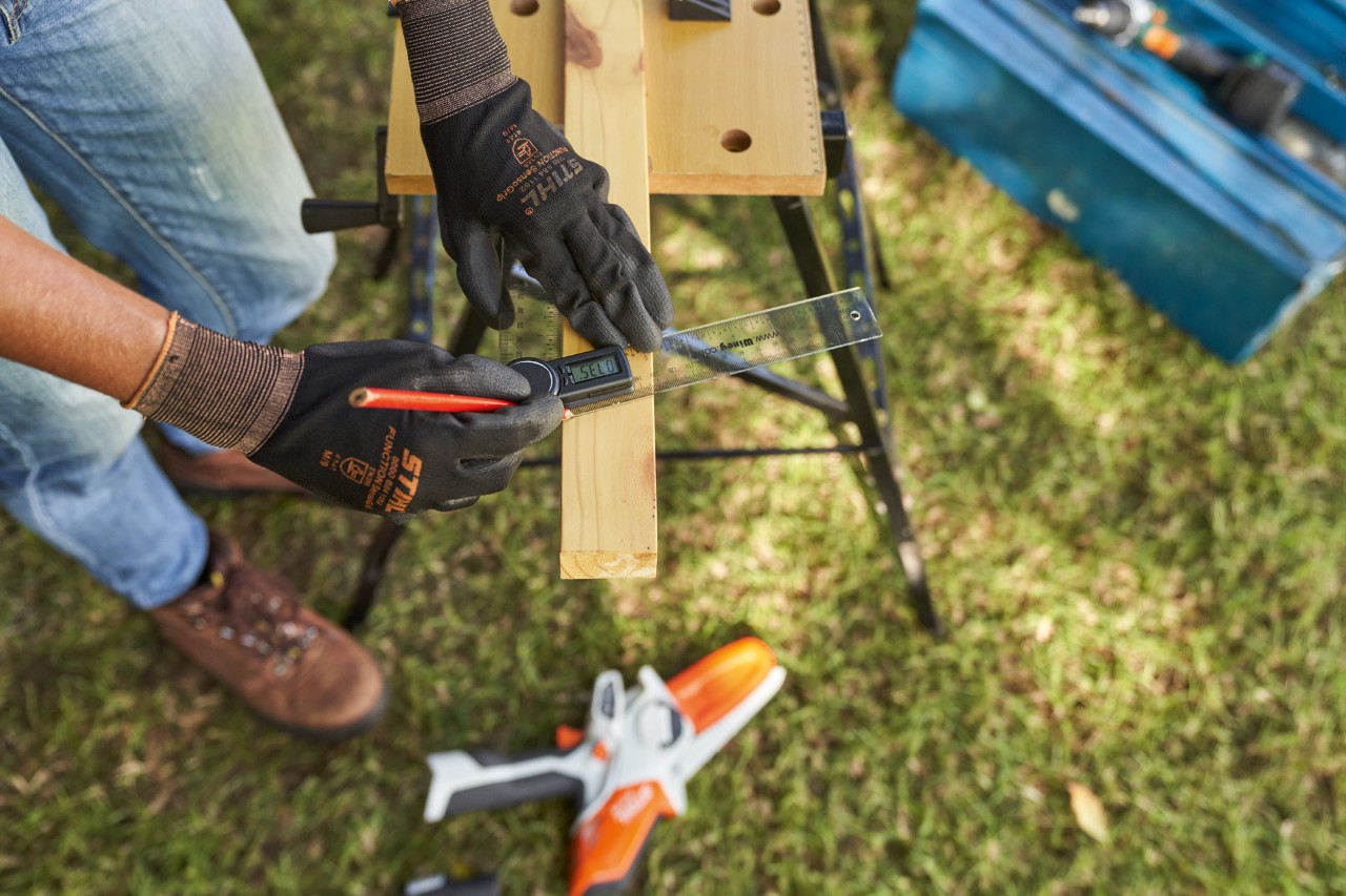Uma mulher com luvas de trabalho calçadas mede a madeira; em baixo, pode ver-se a minipodadora  STIHL GTA 26