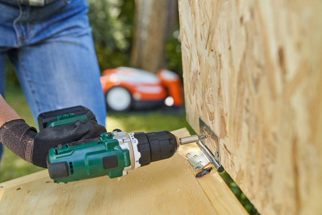 Uma mão com uma luva STIHL FUNCTION SENSOTOUCH aparafusa a dobradiça para o telhado da garagem para robot corta-relva.