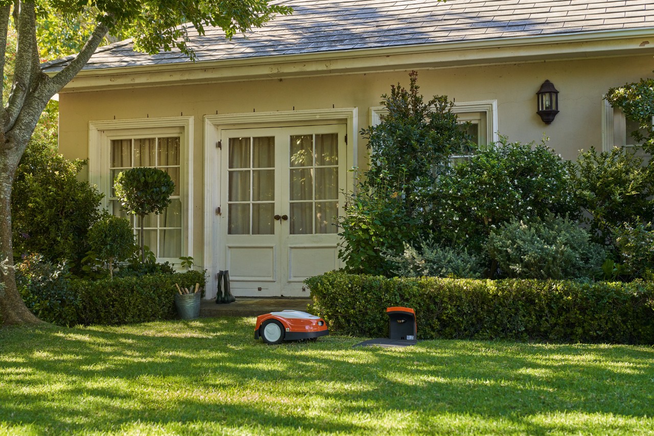 Robot corta-relva STIHL iMOW® junto à estação de carga numa área de relvado verde com uma entrada de casa atrás
