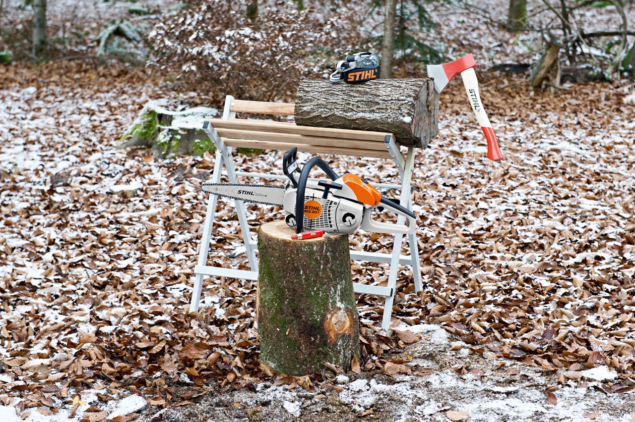 Uma motosserra STIHL MS 201 e um marcador de giz PRO num toro à frente de um cavalete com um toro e um machado AX 6
