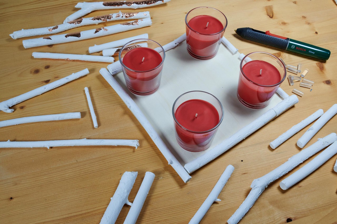 Centro de mesa de Natal de bricolage: quatro velas vermelhas numa tábua de madeira quadrada, rodeada de galhos brancos.