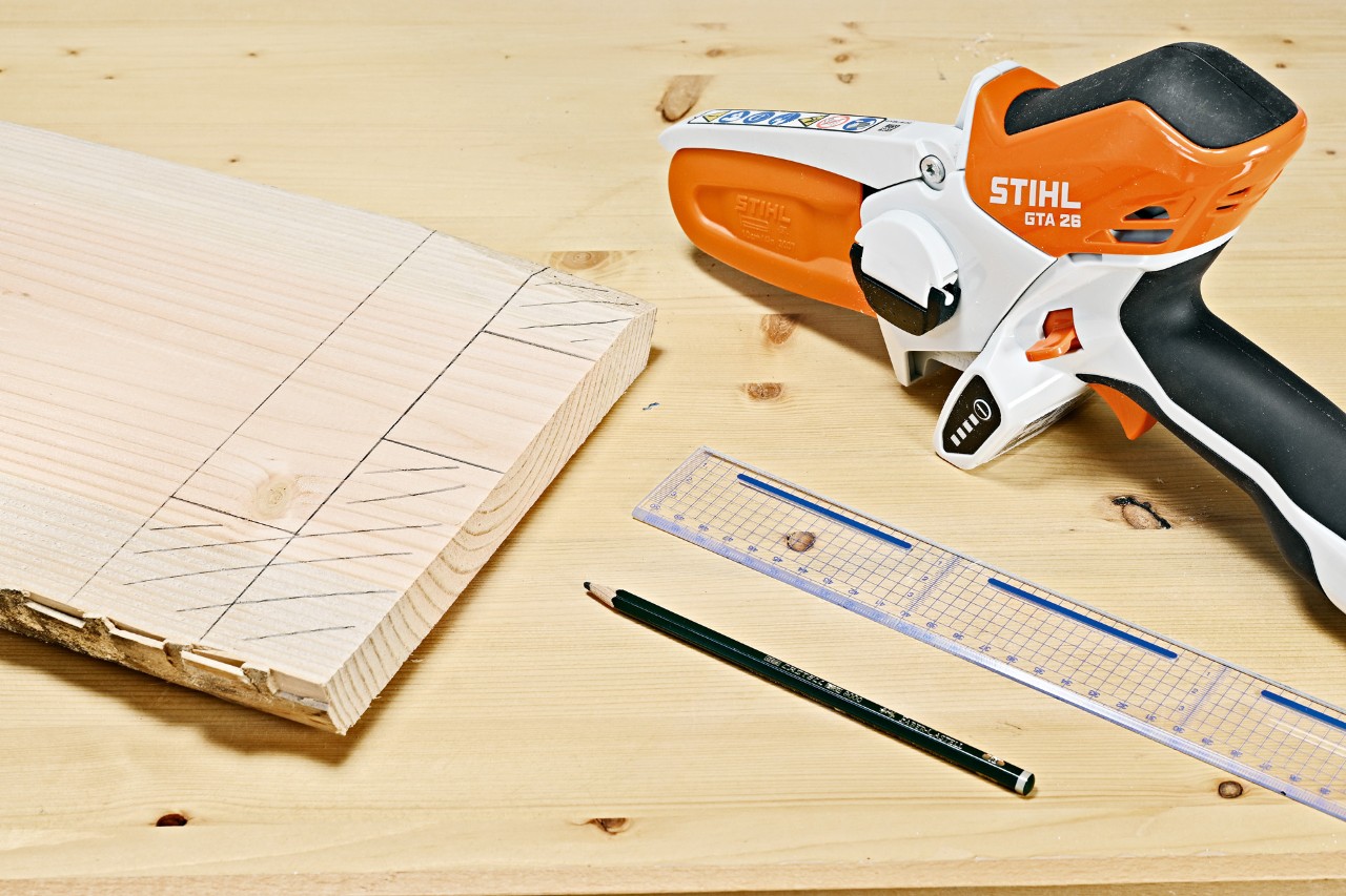 Mesa de apoio de bricolage: traço de lápis numa tábua de madeira ao lado de uma minipodadora STIHL GTA 26, lápis e régua