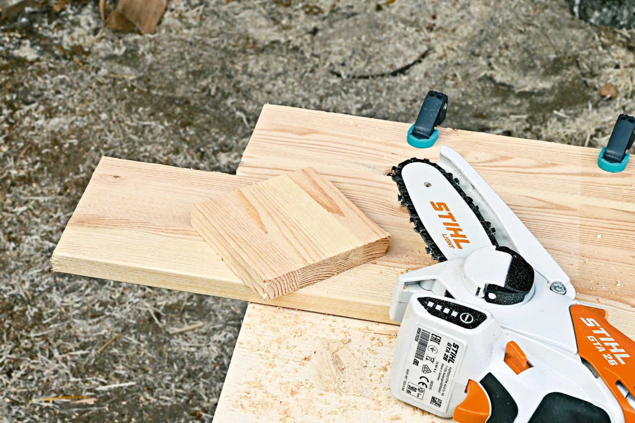 Minipodadora de corrente a bateria STIHL GTA 26 junto de um pedaço de madeira do qual foi cortado um quadrado 
