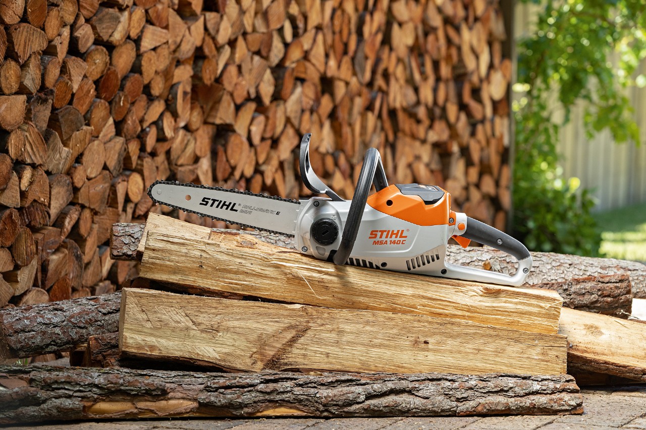A motosserra a bateria STIHL MSA 140 C-B sobre uma pilha de toros compridos, com lenha bem empilhada em segundo plano
