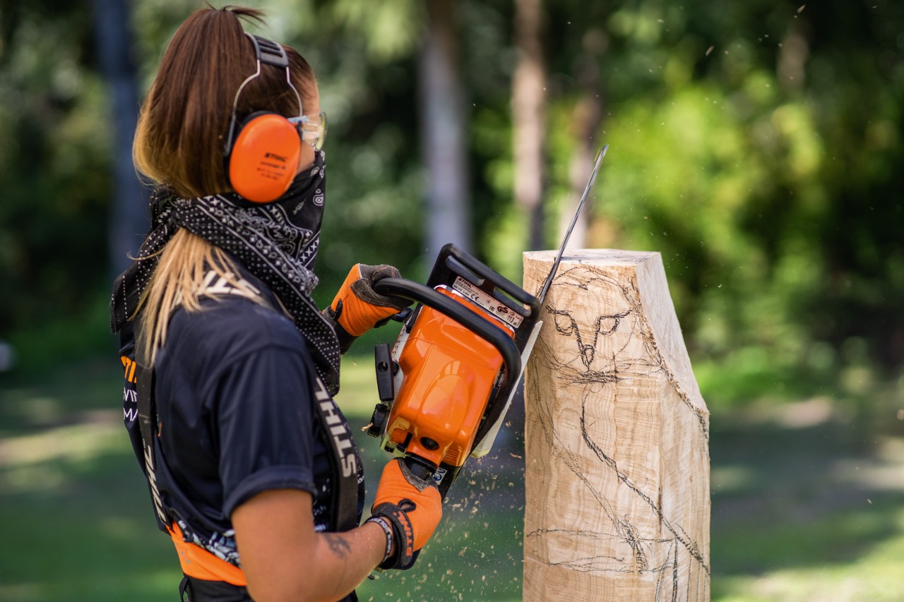 Uma mulher com proteção auditiva, luvas e uma bandana sobre a boca a esculpir uma coruja com a motosserra STIHL.