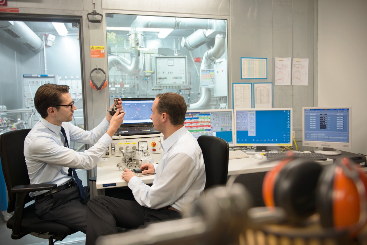 Dois homens sentados num laboratório, numa secretária com várias mini-portas, examinam parte de um novo produto