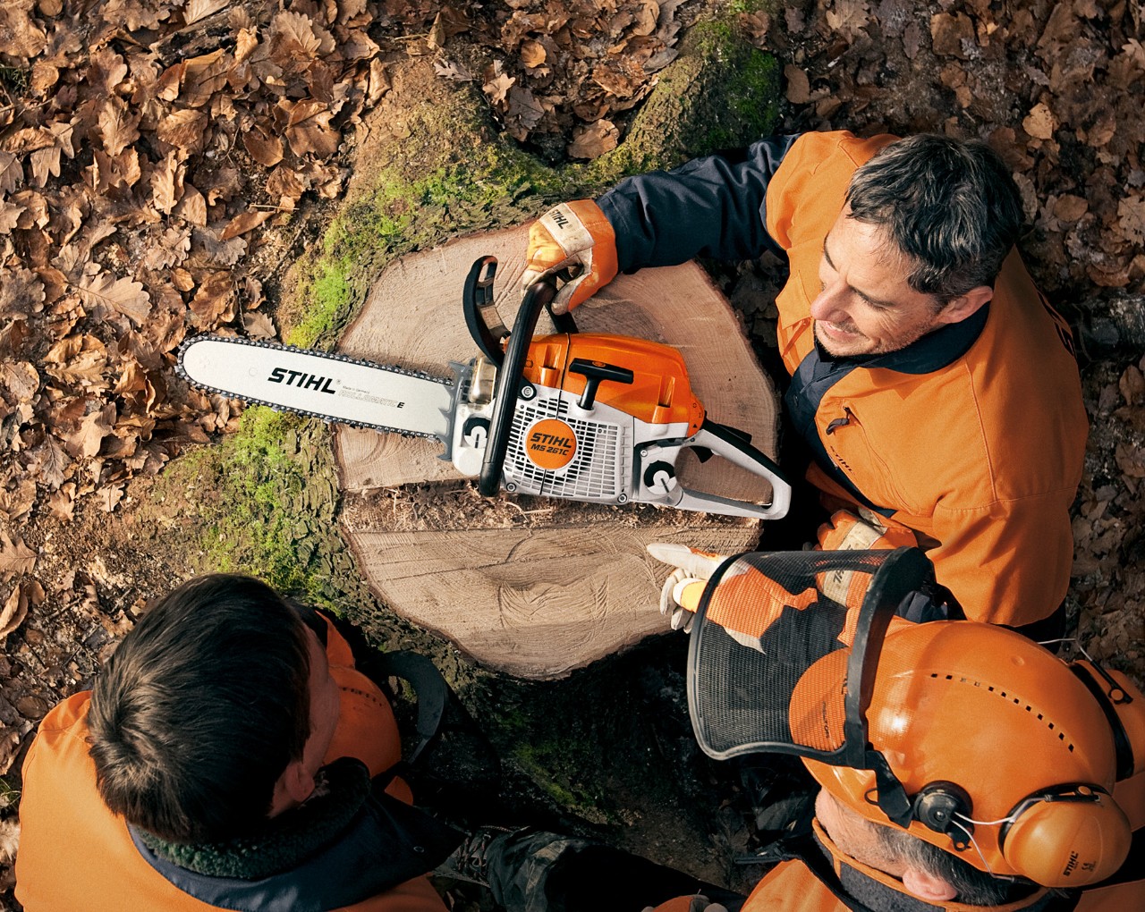 Vista aérea de uma motosserra STIHL MS 261 a gasolina sobre um cepo, com pessoas usando equipamento de proteção à volta