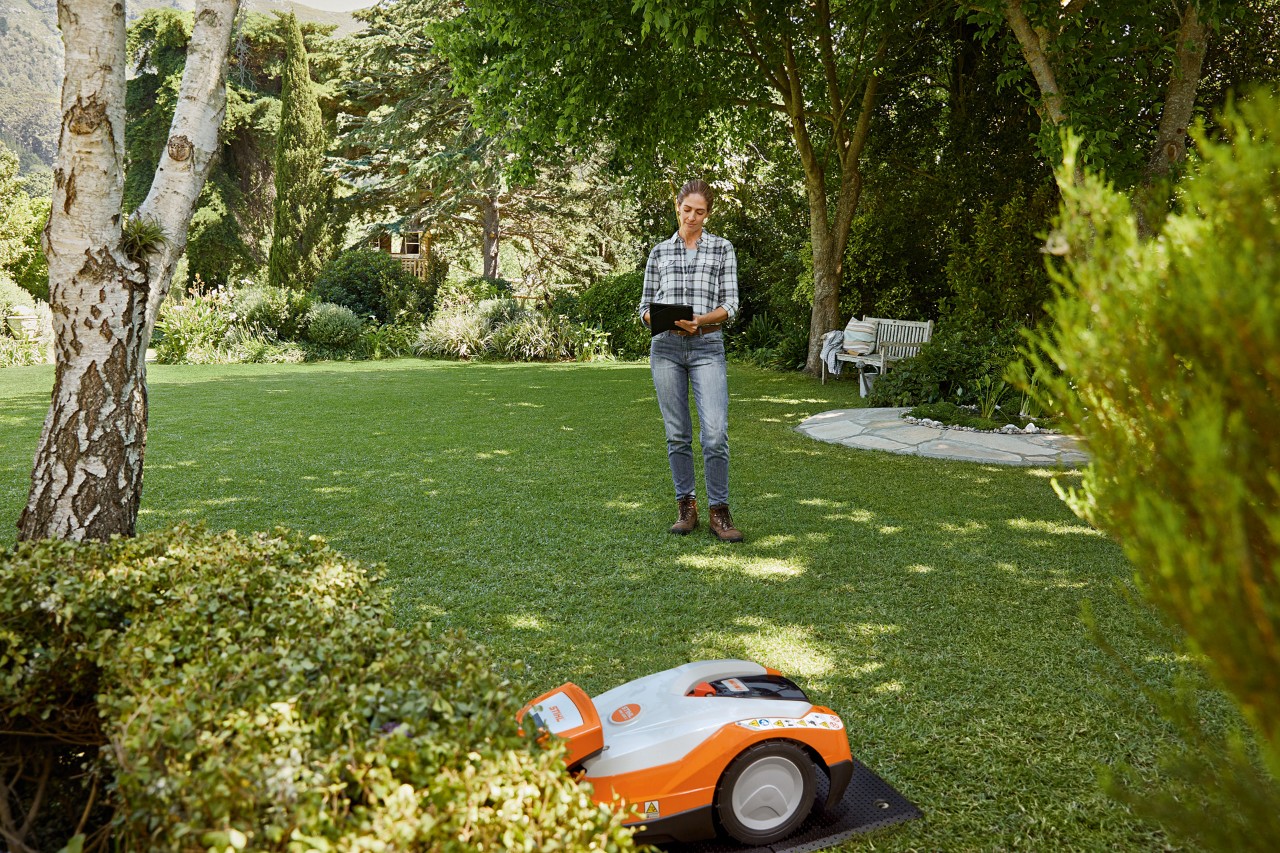 Uma mulher a usar um tablet num jardim e a operar um robot corta-relva STIHL iMOW® 522