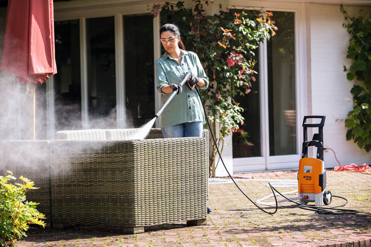 Uma mulher com óculos de proteção está a limpar o mobiliário de jardim no terraço com a máquina de limpeza de alta pressão STIHL RE 88