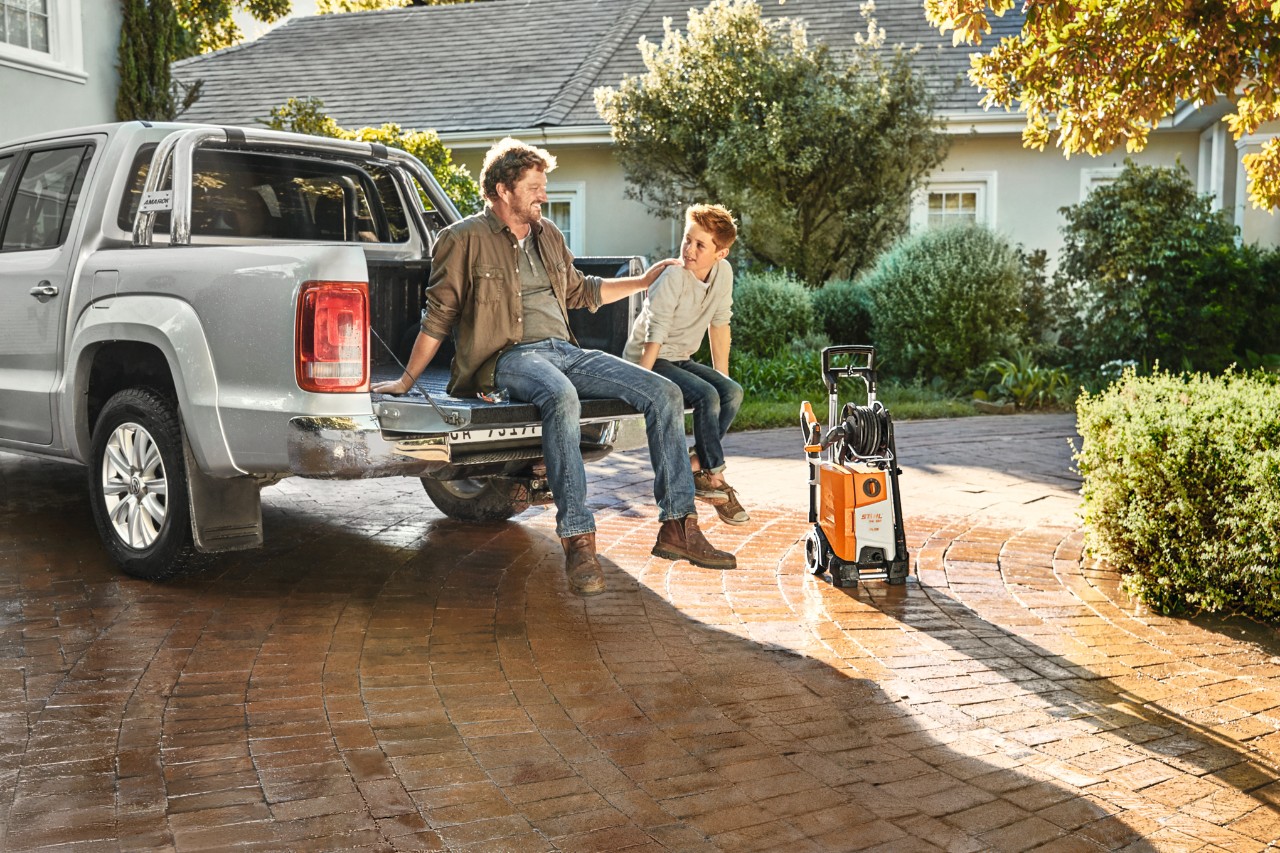 Pai e filho sentados na superfície de carga de uma pickup e ao lado do veículo está uma máquina de limpeza de alta pressão STIHL RE 130 plus